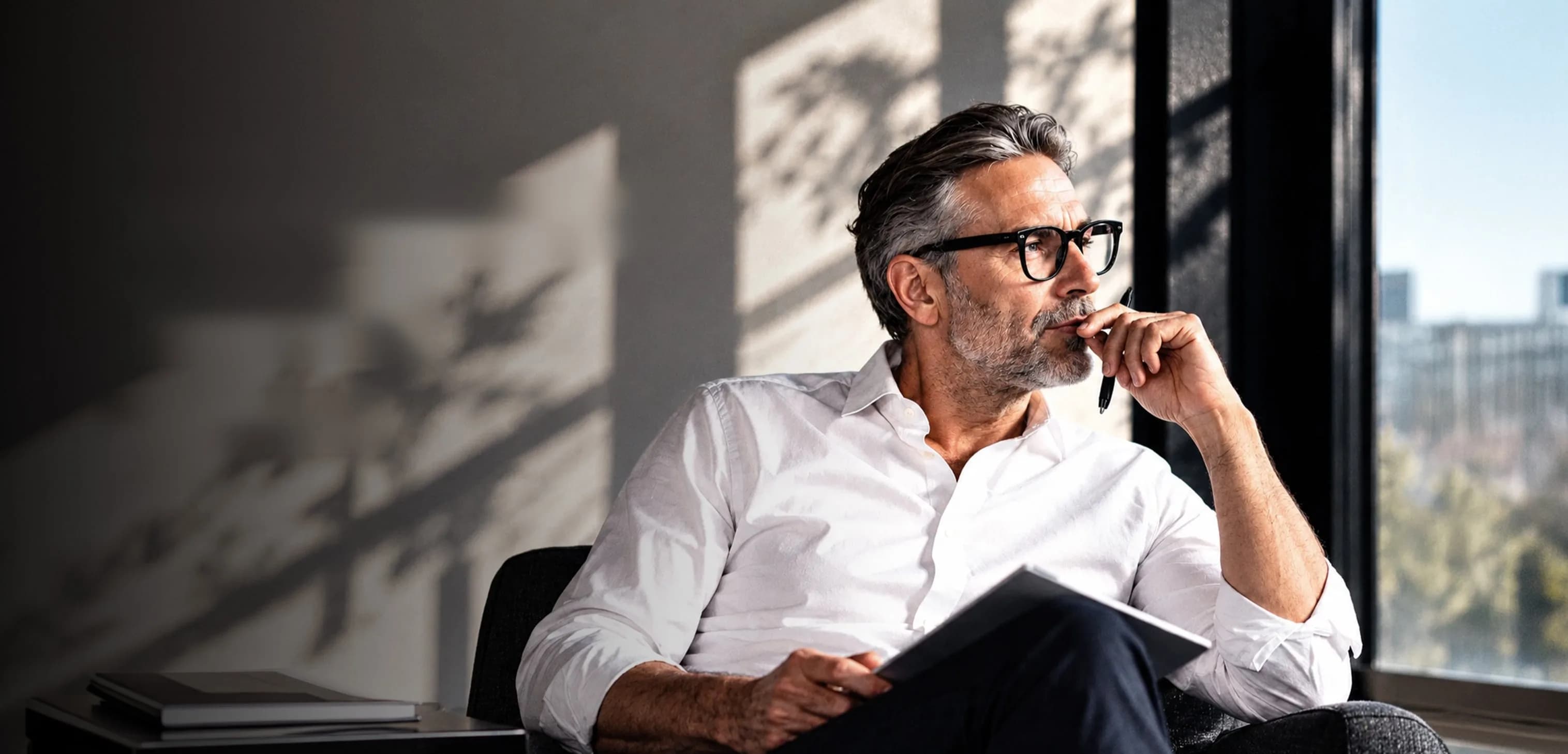 A professional pausing in thought by an office window, a notebook resting on their lap